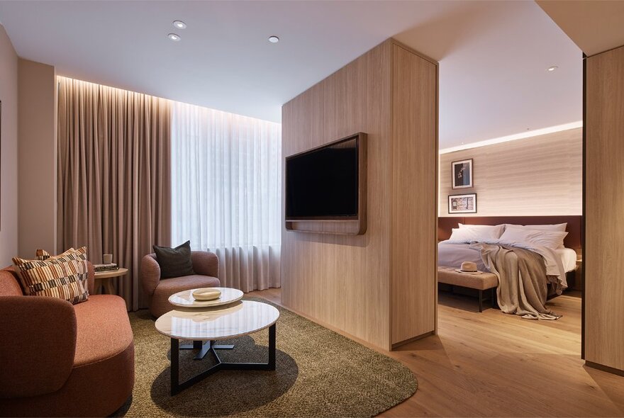 Hotel suite with a bedroom and lounge area separated by a wall with large TV screen.