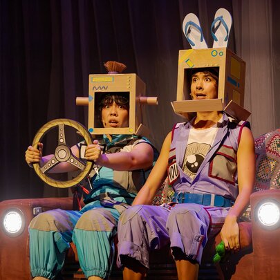 Two people sitting on a couch with headlights on stage, wearing cardboard helmets and one holding a cardboard steering wheel.