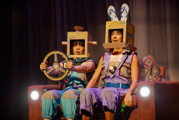 Two people sitting on a couch with headlights on stage, wearing cardboard helmets and one holding a cardboard steering wheel.