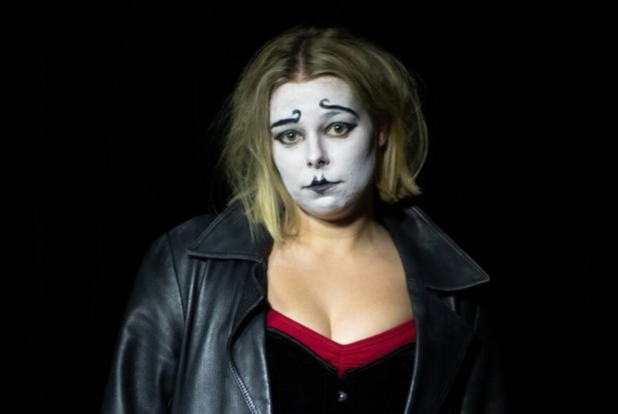 A performer from the cabaret, dressed in a black leather coat, with white theatrical face makeup and exaggerated lips and eyebrows.