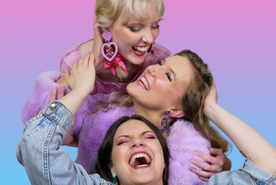 Three women hugging and smiling vertically, with pink and blue background.