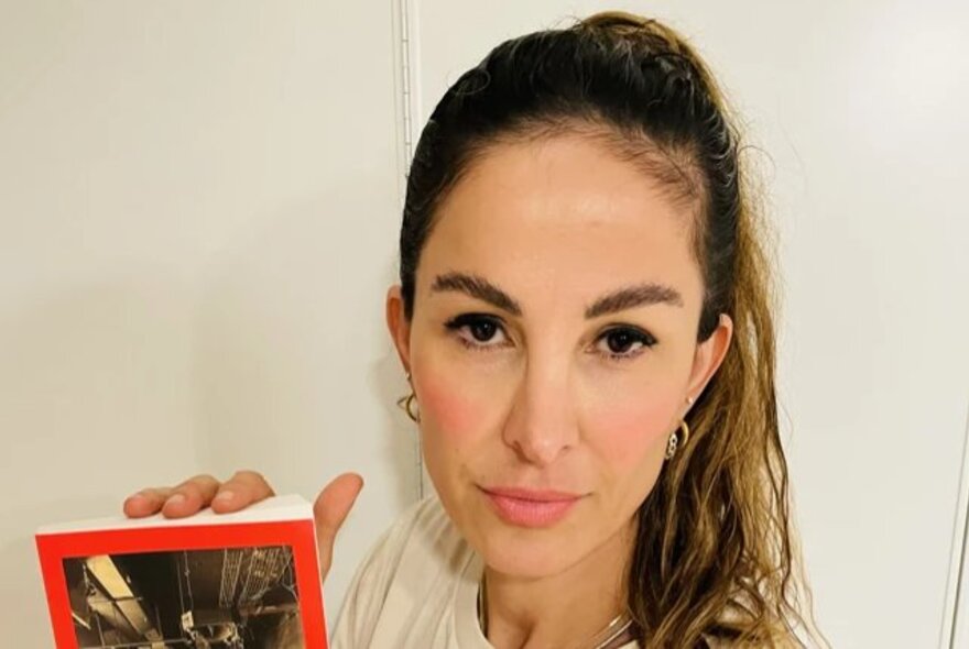 Dr Saya Aziz looking directly at the camera with a neutral expression, her brown hair pulled back into a high ponytail, the corner of a book she holds is visible in the left corner of the frame. 