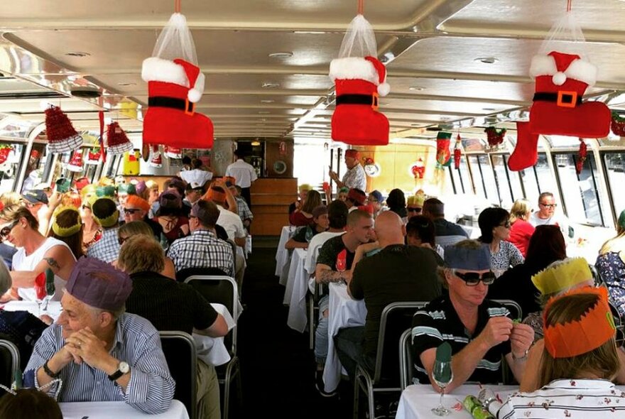 People wearing party hats seated at tables inside a cruise boat cabin decorated with Santa boots and other Christmas decorations.