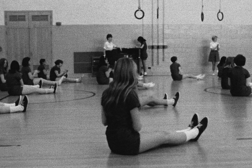 A black and white image of a gym class with girls seated on the floor with legs out front. 