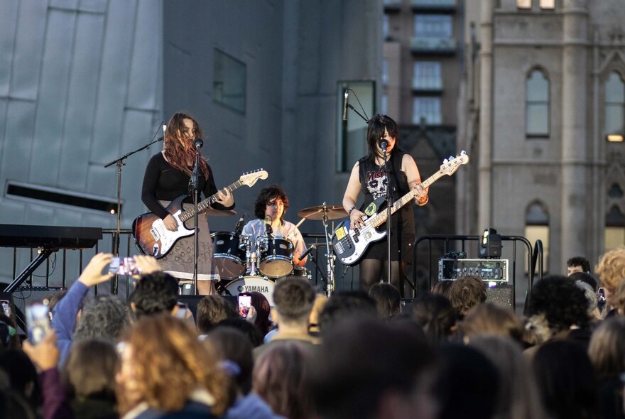 A young band on stage at Fed Square, with two guitarists and a drummer.