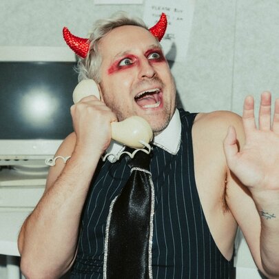 Man wearing devil's horns and make-up, holding a phone in front of a computer, his hands in the air and mouth open.