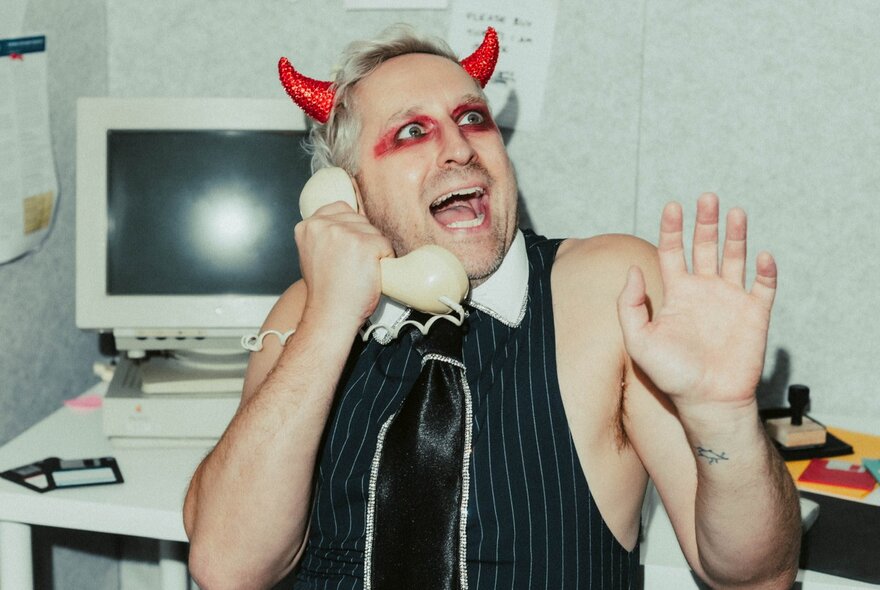 Man wearing devil's horns and make-up, holding a phone in front of a computer, his hands in the air and mouth open.