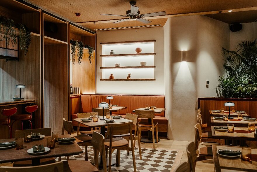 Interior of a restaurant and bar with warm lamp lighting on the wall, arrangements of small tables and chairs, and a patterned black and white check floor.