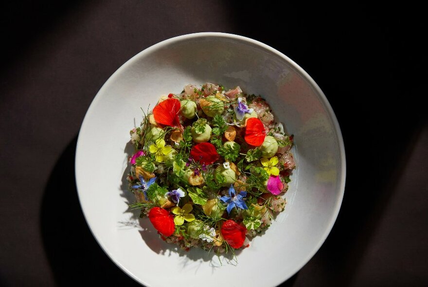 Very colourful arrangement of food and flower petals in a white bowl.