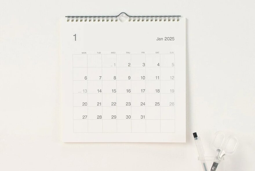 Blank calendar hanging on a wall with a small pen cup next to it storing a pen and scissors.