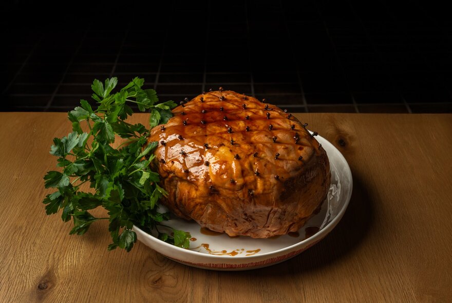 A Christmas glazed ham in a round serving bowl, a large sprig of parsley as garnish.