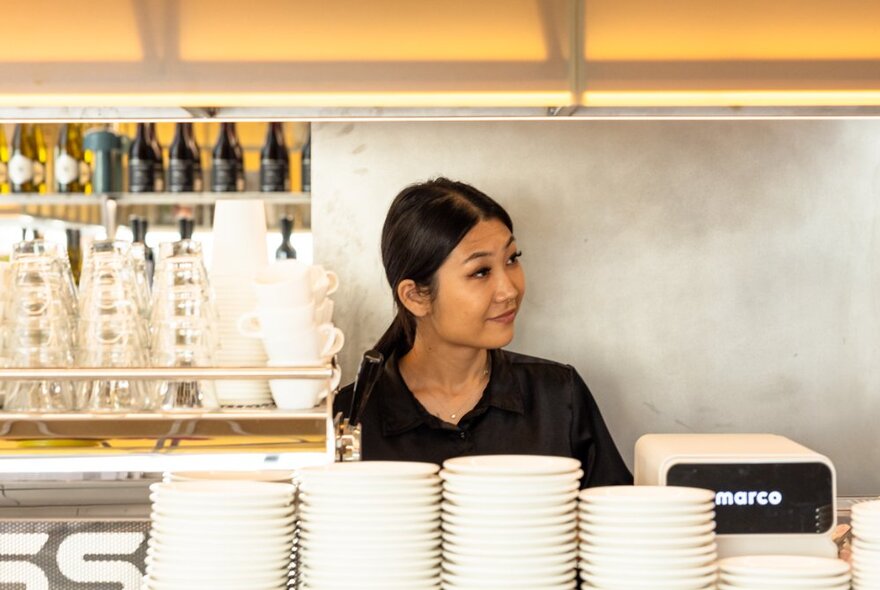 A person standing behind the coffee machine inside a cafe, stacks of white saucers in front of her.