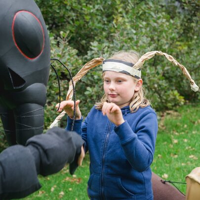 An adult performer dressed in a black ant costume, interacting with a child wearing a headband with ant antennae, in a grassy outdoor space.