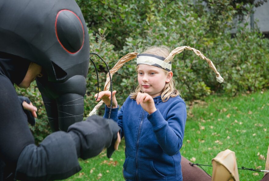 An adult performer dressed in a black ant costume, interacting with a child wearing a headband with ant antennae, in a grassy outdoor space.