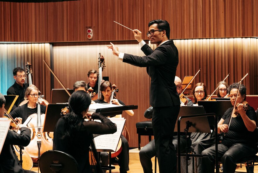 A man in a black suit conducting members of Orchestra Victoria as they play their musical instruments in a hall.