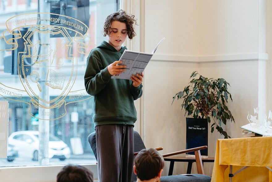 A young teenager standing up in front a group reading from a notebook, in front of a large window.