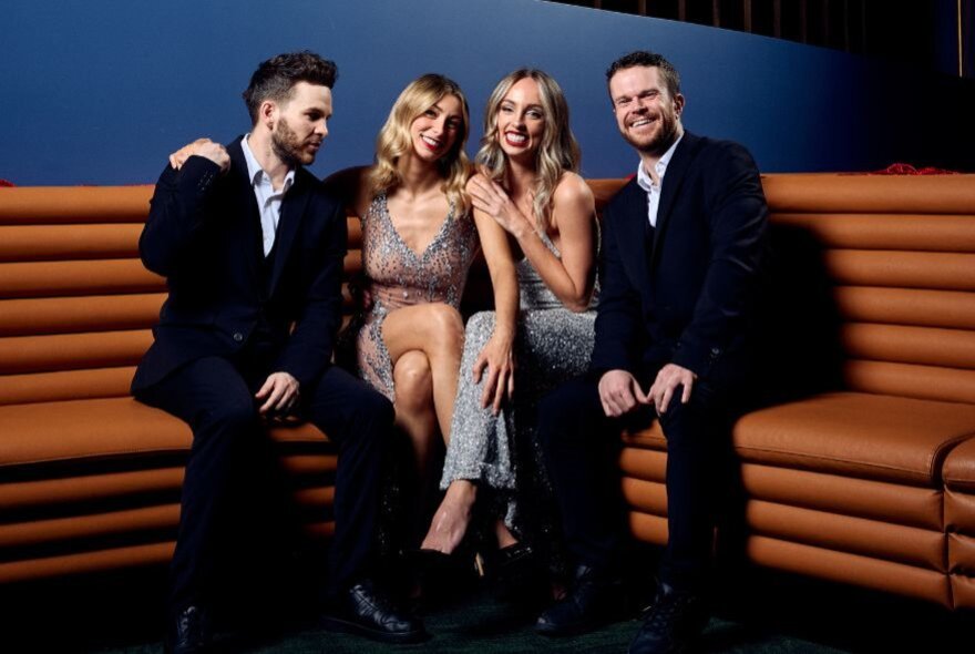 A group of smiling people, two men and two women, on a tan sofa in smart clothing.
