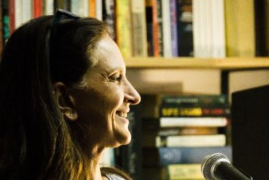 Profile view of poet Indrani Perera, smiling while speaking into a microphone with bookshelves in the background.