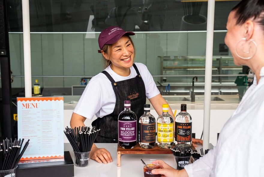 A barista behind a counter with bottles of coffee flavourings displayed in front of her, talking to a customer in a white shirt.