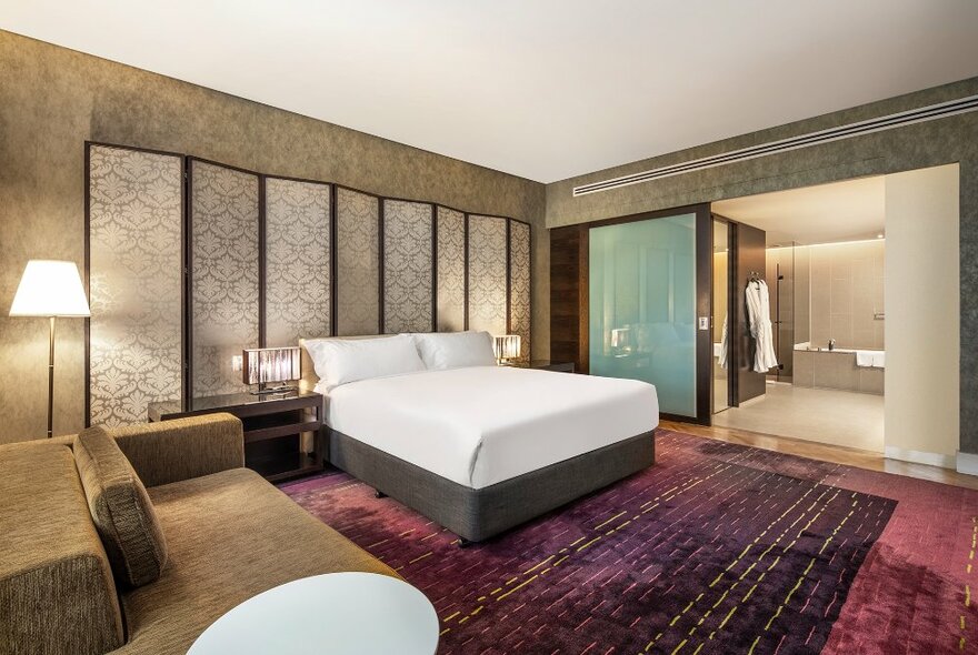 A suite at InterContinental Melbourne Rialto with a king-sized bed, a large couch, rugs on a carpeted floor and a view into the bathroom in the background. 