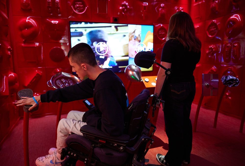 Two people exploring an interactive red room in gallery. One is sitting in a wheelchair and pushing a button.