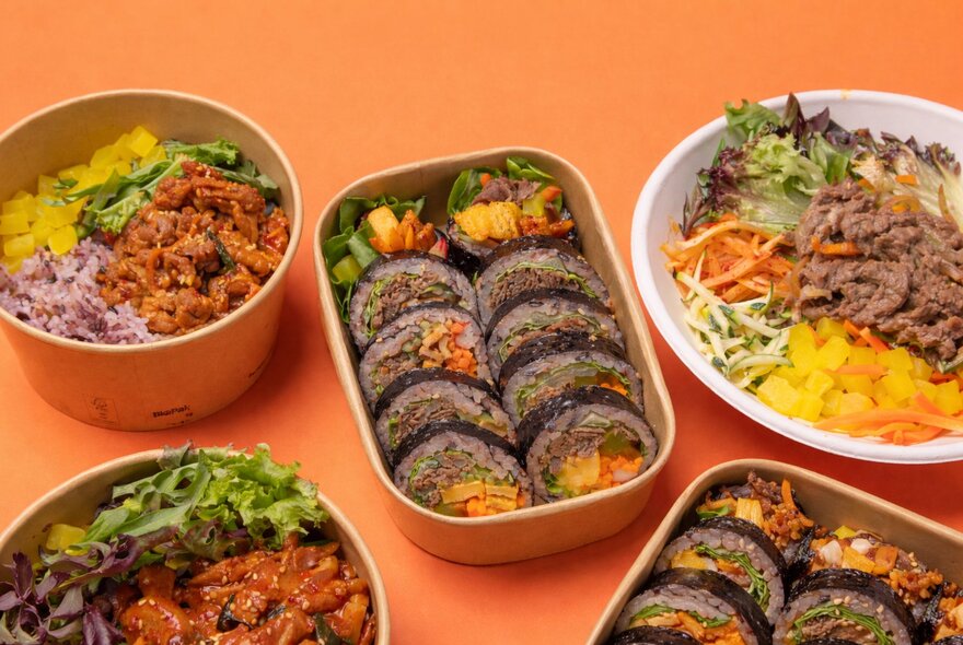 A selection of Korean dishes in bowls and plates on an orange coloured table.