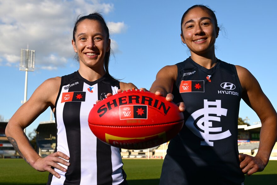 Carlton and Collingwood AFLW captains smiling on a football ground and holding forward at Sherrin football. 