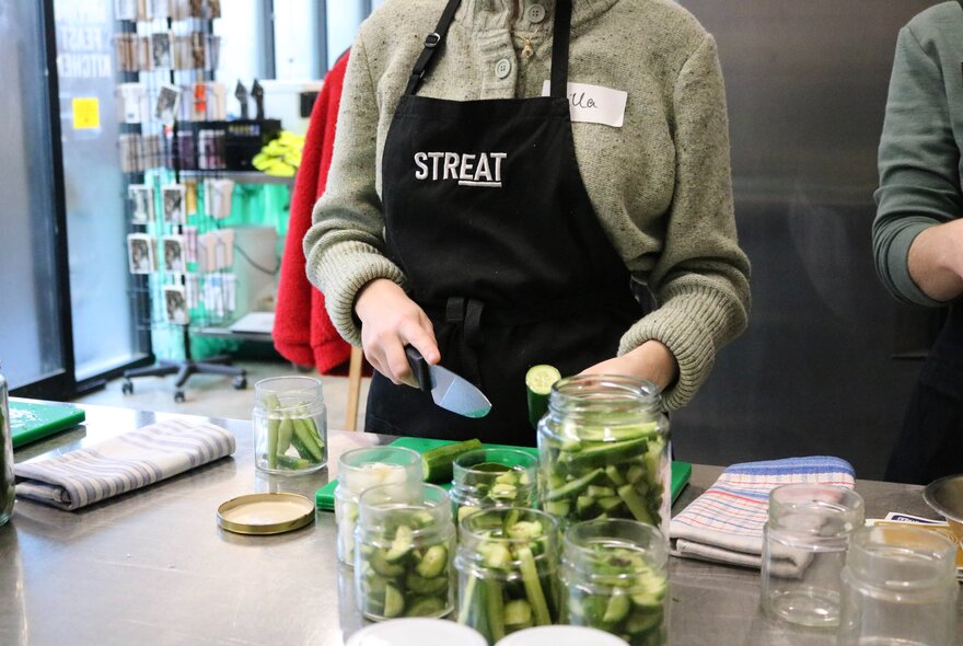 Person wearing an apron holding a sliced cucumber next to sliced vegetables in pickling jars.