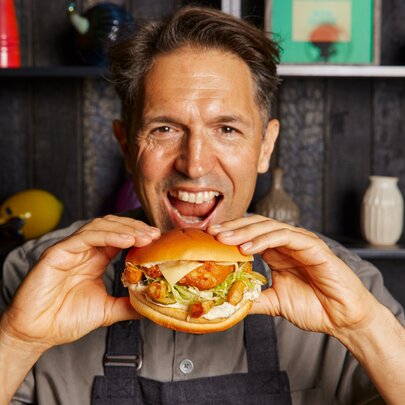 Chef Ben Shewry lifting a fish burger to his mouth, smiling.