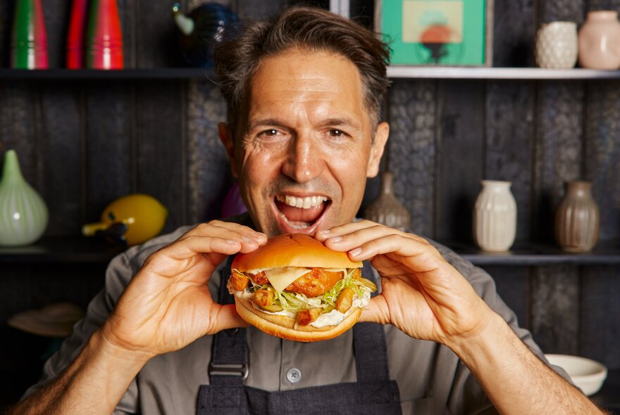 Chef Ben Shewry lifting a fish burger to his mouth, smiling.
