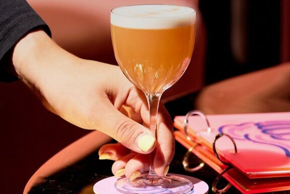 Hand placing a pink cocktail on a table next to a book.