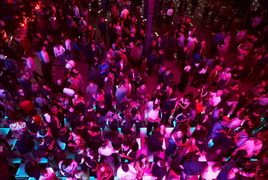 Elevated, overhead view of a crowded dance floor with a large group of people dancing under vibrant pink and purple lighting.