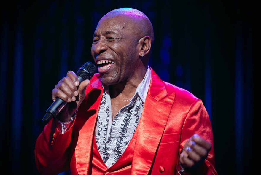 Bald singer performing with a microphone, wearing a shiny red suit and patterned black and white shirt.