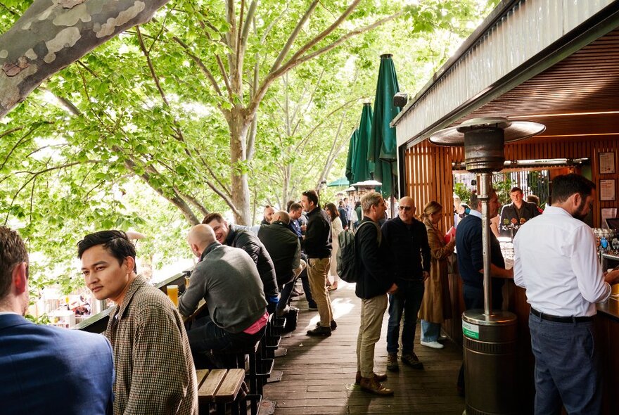 People seated and standing on the outdoor deck at Arbory Bar and Eatery, beside the Yarra River, with leafy green trees in the background.