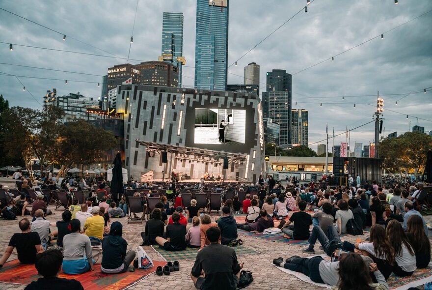 A crowd of people seated on picnic rugs and deck chairs outside at Fed Square, watching an open air film on a large screen.
