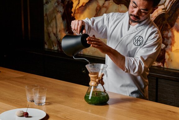 Japanese chef pouring matcha into a jug at a wooden counter.