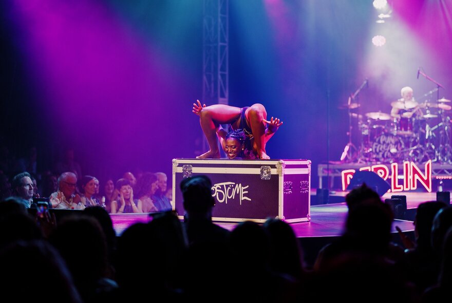 A cabaret contortionist standing on a box on stage in a deep backward bend with her head between her feet, before a seated audience; purple lights on her.