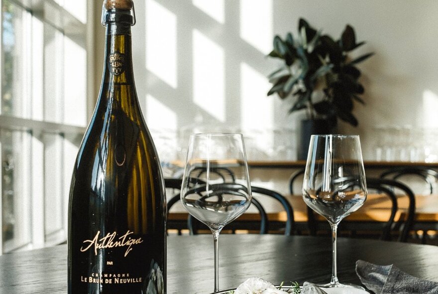 A bottle of white wine next to two empty wine glasses on a dark timber table, windows in the background.