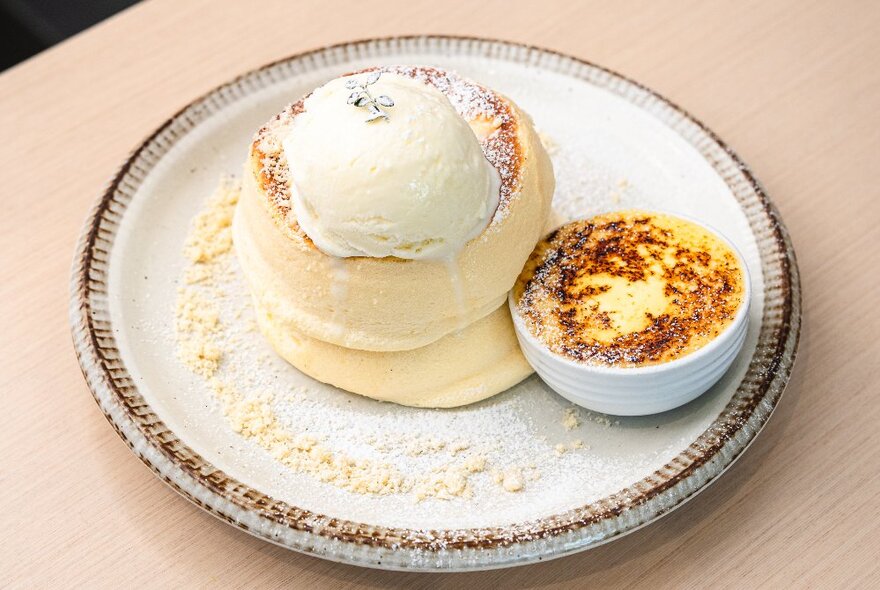 Souffle-style fluffy pancakes with a dollop of vanilla ice cream and a creme brulee in a small white bowl beside it.