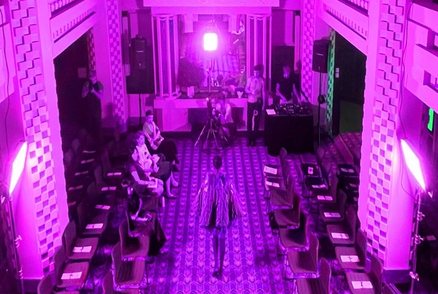 A person walks down a purple-lit aisle in a venue, with seating on both sides and a stage area in the background.