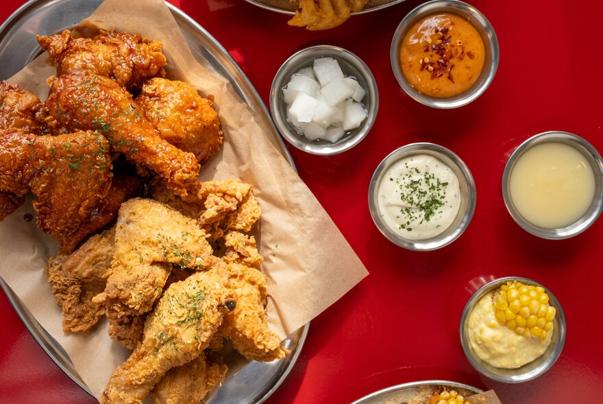 A silver platter with brown paper on top filled with Korean fried chicken, next to an assortment of small silver bowls with different dipping sauces.