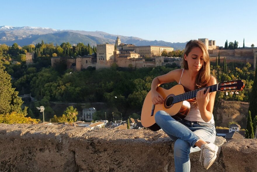 Laura de Elvira playing acoustic guitar outdoors with Spanish scenery in the background.