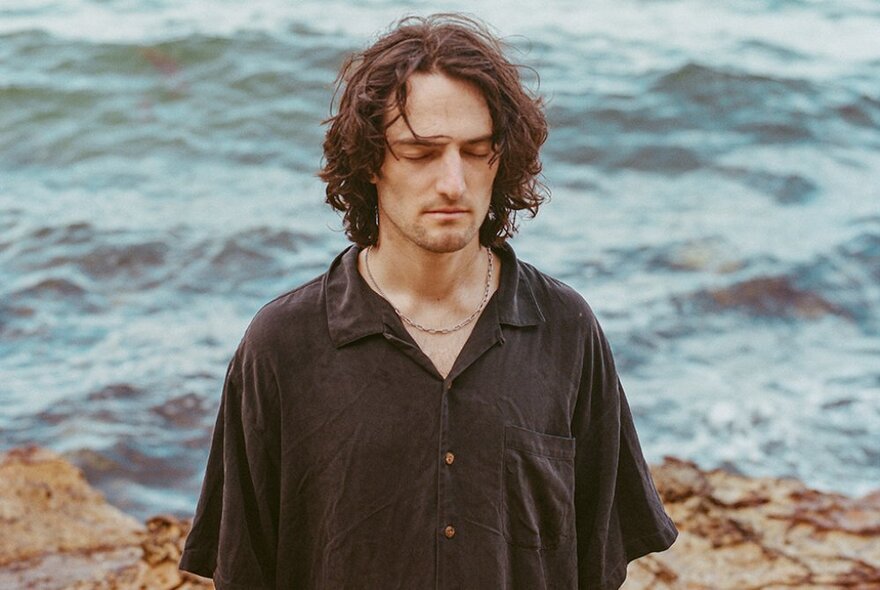 A downcast man wearing a loose brown shirt on rocks in front of the ocean.