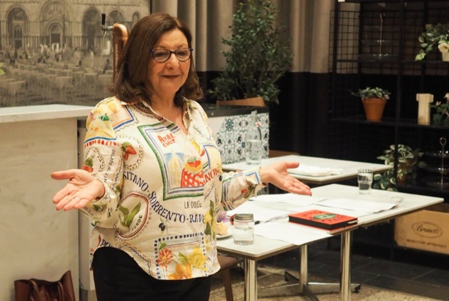 Tour leader speaking with her arms open to the side, tables in the background.
