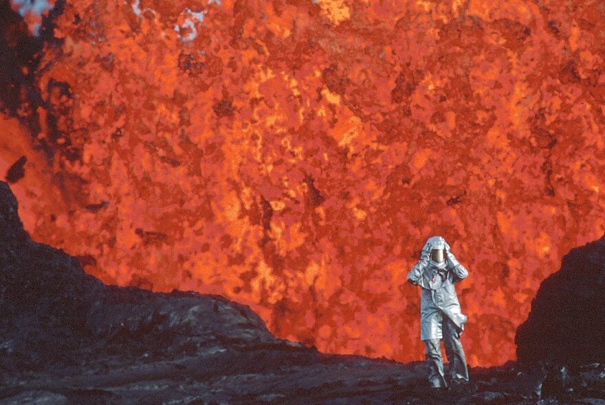 Film still of a small figure in a silver astronaut outfit and helmet, standing next to a volcano that is spewing out orange lava.