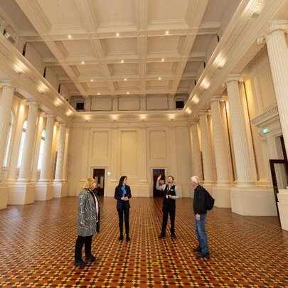People on a small tour at the Immigration Museum, in an empty room with columns and a tiled floor.
