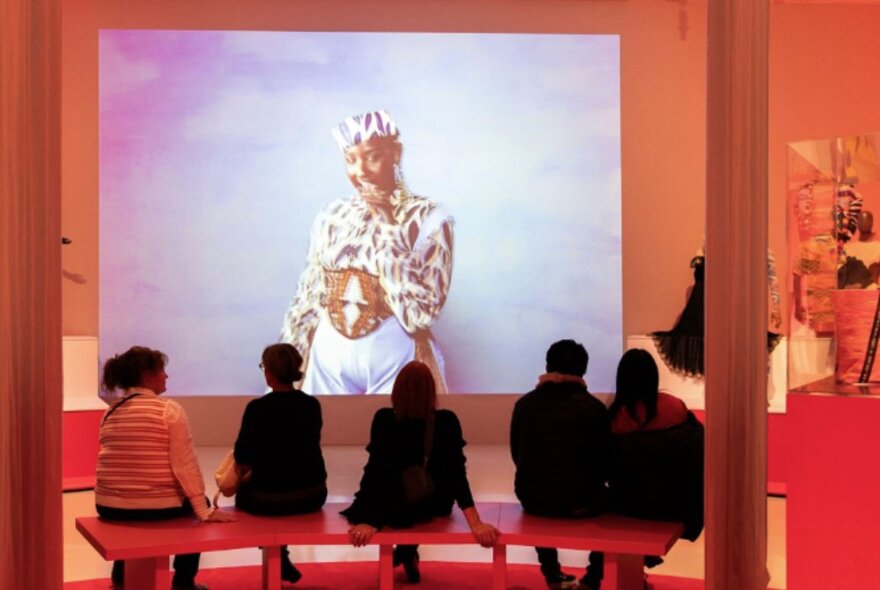 People sitting on a bench seat at the NGV, looking at an image projected onto the gallery wall.