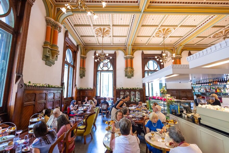Customers sitting in a high tea restaurant with a mix of marble and wooden antique furniture, decorative half wooden walls and extravagant high ceilings with chandeliers.