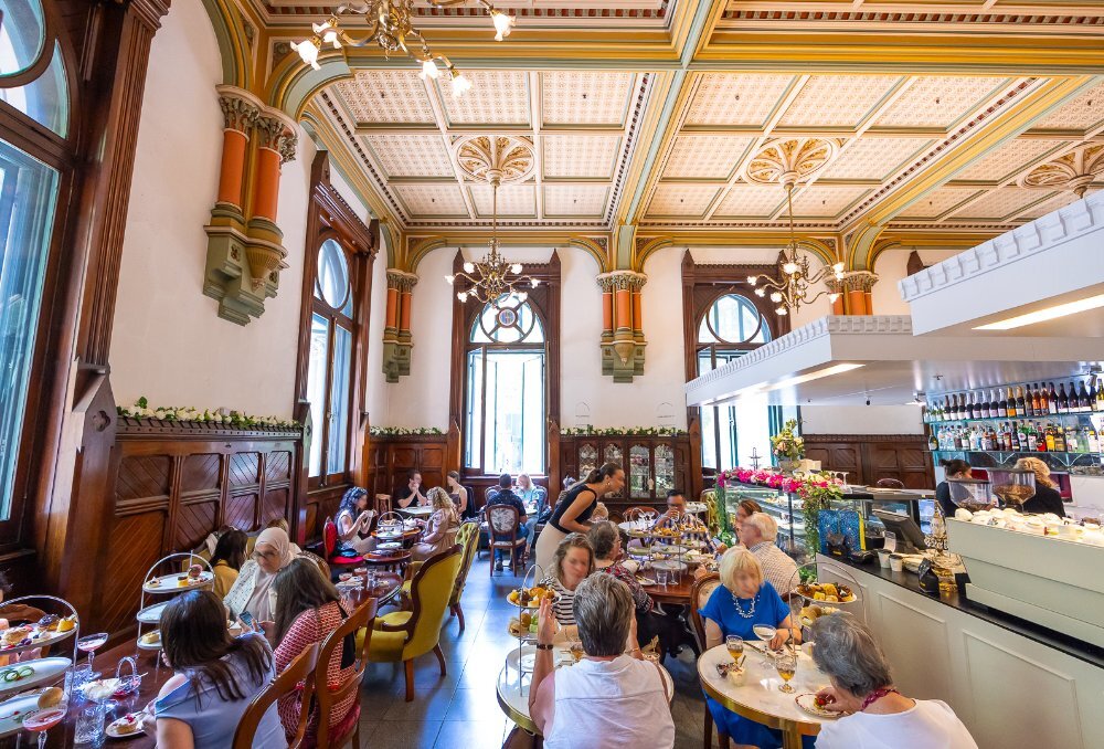 Customers sitting in a high tea restaurant with a mix of marble and wooden antique furniture, decorative half wooden walls and extravagant high ceilings with chandeliers.