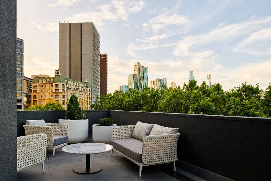 A room's balcony at The Royce Hotel, with an outdoor couch and table; a view of the evening skyline with tall buildings and trees visible against a golden sky.